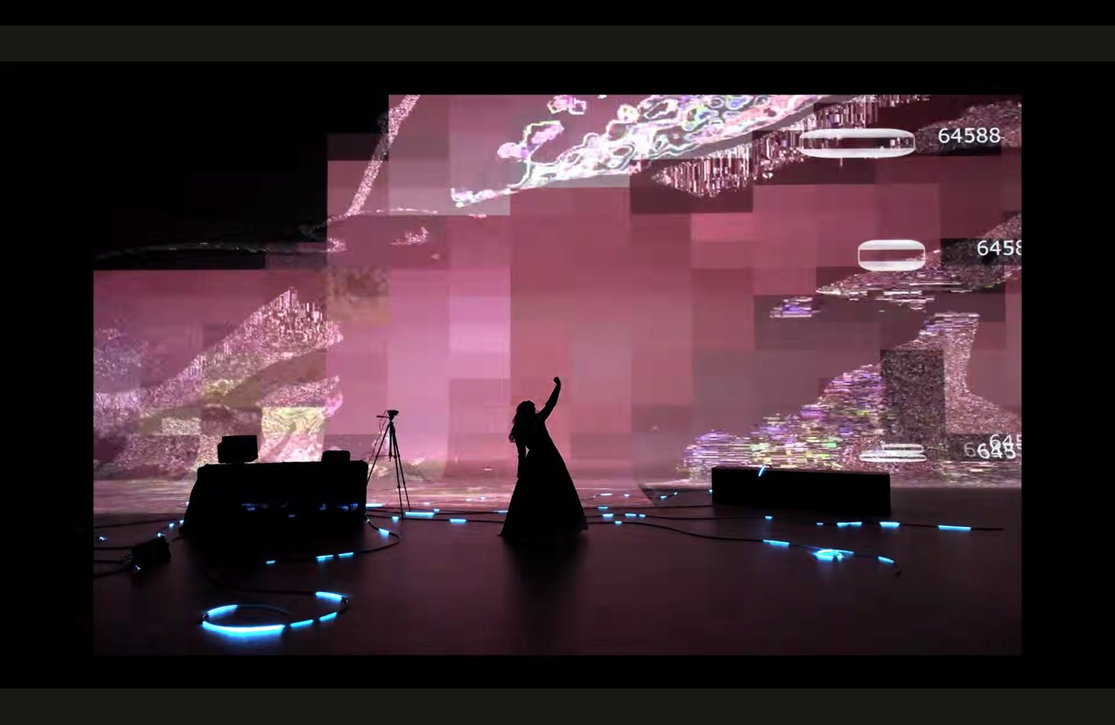 Het silhouet van een performer in een lange jurk steekt een arm omhoog voor een grote roze glitchprojectie; blauwe lichtlijnen liggen op de vloer.
