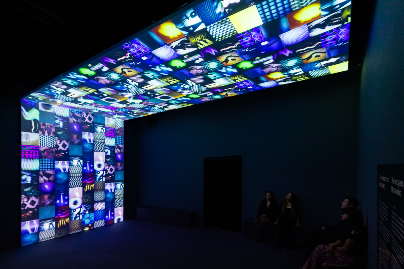 Een donkere zaal waarvan het plafond en een wand volledig bestaan uit honderden videoschermen; bezoekers zitten op een bank en kijken omhoog.